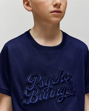A child wears the Psycho Bunny KIDS ELIAS GRAPHIC TEE (KT0200013), a navy Pima cotton shirt featuring an embroidered "Psycho Bunny" text and a small Bunny graphic skull logo on the front; image cropped at shoulders and chin.