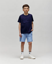 A boy with short brown hair and a neutral expression stands against a plain background, wearing the Psycho Bunny KIDS ELIAS GRAPHIC TEE (KT0200013) in navy, light blue shorts, white socks, and white sneakers, with one hand in his pocket.