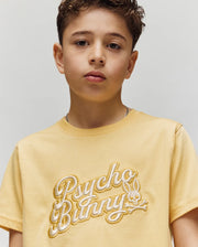 A young boy with short, curly brown hair wears the Psycho Bunny KIDS ELIAS GRAPHIC TEE (KT0200013), a yellow Pima cotton shirt with a Bunny graphic and "Psycho Bunny" logo, standing against a plain light background, looking slightly serious.