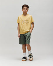 A boy with short curly hair and a neutral expression wears Psycho Bunny's KIDS ELIAS GRAPHIC TEE (KT0200013) in retro yellow, paired with green shorts, white socks, and black-and-white sneakers against a plain background.