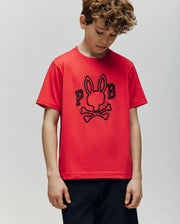 A young boy with curly hair wears the Psycho Bunny KIDS BALES GRAPHIC TEE (KT0200021), a red Pima cotton top featuring a Big Bunny print and "P B" letters, paired with black shorts. A plain background highlights its year-round versatility.