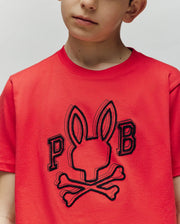 A child wears the Psycho Bunny KIDS BALES GRAPHIC TEE (KT0200021), a red Pima cotton tee with the signature Big Bunny graphic above crossbones and "P" & "B" accents—a perfect year-round staple.