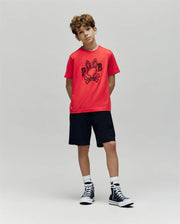 A young boy with curly hair stands against a gray background, wearing the Psycho Bunny KIDS BALES GRAPHIC TEE (KT0200021) in red with a Big Bunny print, black shorts, white socks, and black high-tops—ideal for year-round style.