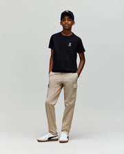 A boy stands against a light gray background, wearing the Psycho Bunny KIDS MISCHIEF BUNNY TEE (KT0200023) in black with an embroidered bunny on the chest, paired with a black cap, beige cargo pants, and white sneakers with black accents.