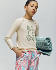 A young boy wears the Psycho Bunny KIDS LONG SLEEVE RASHGUARD - KT0200027 with UPF 50+ UV protection, bunny-patterned shorts, and holds a green towel under his arm against a plain background.