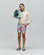 A young person holds folded towels, wearing the Psycho Bunny KIDS LONG SLEEVE RASHGUARD - KT0200027 with a beige bunny print and UPF 50+ protection, patterned bunny shorts, and blue slides against a plain backdrop.
