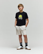 A boy stands with hands in pockets, wearing a Psycho Bunny KIDS PETE TENNIS GRAPHIC TEE (KT0200038) in black, made from soft Pima cotton, along with cream shorts, black patterned socks, and white sneakers against a light gray background.