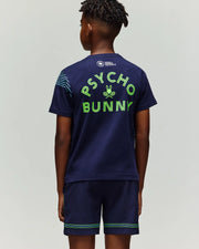 A boy faces away wearing the Psycho Bunny RETAIL BALL CREW YOUTH TEE (FR) - KT0200042, a navy blue t-shirt with "PSYCHO BUNNY" in bright green letters and a bunny with crossed racquets graphic below.