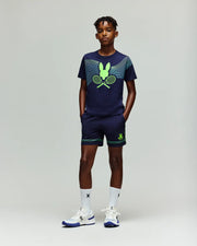 A young boy wears the Psycho Bunny RETAIL BALL CREW YOUTH TEE (FR) - KT0200042, a navy blue sports shirt with green accents, bunny logo and crossed tennis rackets, paired with white socks and sneakers against a plain background.