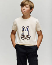 A boy with light brown hair wears the cream KIDS WADE GRAPHIC TEE - KT0200054 by Psycho Bunny, featuring an embroidered Bunny and crossbones. Made from 100% cotton, he pairs it with black pants and stands against a plain background.