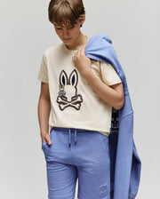 A child wears the KIDS WADE GRAPHIC TEE - KT0200054 by Psycho Bunny, a beige 100% cotton tee with an embroidered Bunny and crossbones, paired with blue shorts and a matching blue sweatshirt draped over one shoulder.