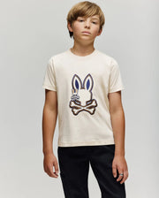 A boy with light brown hair wears the Psycho Bunny KIDS WADE GRAPHIC TEE - KT0200054, a cream 100% cotton shirt featuring an embroidered Bunny and crossbones graphic, standing neutrally against a plain light background.