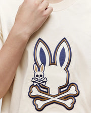 Someone wearing the Psycho Bunny KIDS WADE GRAPHIC TEE—KT0200054, a cream-colored 100% cotton shirt with a large embroidered Bunny head above crossed bones and a smaller bunny skull and crossbones on the left side.