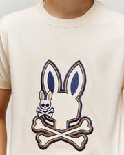 A person wearing the Psycho Bunny KIDS WADE GRAPHIC TEE (KT0200054), a cream 100% cotton shirt with a large bunny head above crossed bones graphic and a small embroidered Bunny with skull and crossbones.