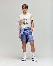 A teenage boy stands against a plain background wearing the Psycho Bunny KIDS WADE GRAPHIC TEE - KT0200054 in beige with an embroidered bunny, paired with light blue shorts, a matching sweatshirt tied around his waist, white socks, and Nike sneakers.