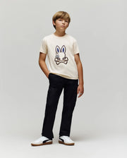 A boy poses against a plain background, wearing the Psycho Bunny KIDS WADE GRAPHIC TEE - KT0200054 in cream, made from 100% cotton with an embroidered Bunny and crossed bones, paired with black pants and white sneakers.