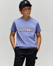 A boy in a black cap, black pants, and the Psycho Bunny KIDS WADE GRAPHIC TEE (KT0200055)—a lavender 100% cotton shirt featuring "PSYBUNY" and a bunny skull logo—stands with hands in pockets against a plain light gray background.