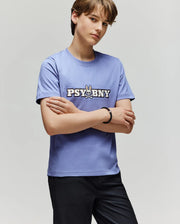 A person with short brown hair stands with arms crossed, wearing the 100% cotton Psycho Bunny KIDS WADE GRAPHIC TEE (KT0200055) in light purple, featuring "PSYBNY" and a bunny graphic, paired with black pants against a plain background.