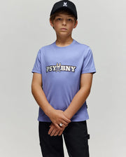 A young boy in a black cap and Psycho Bunny KIDS WADE GRAPHIC TEE (KT0200055) stands against a light gray background, looking at the camera with hands clasped. The lavender tee features 100% cotton, regular fit, cartoon bunny, and "PSYBNY" text.