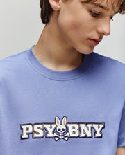 A child wears the Psycho Bunny KIDS WADE GRAPHIC TEE (KT0200055) in light blue—100% cotton, regular fit—featuring "PSYBNY" and a bunny-eared skull and crossbones, looking down and slightly to the side.