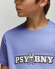 A young person wears the Psycho Bunny KIDS WADE GRAPHIC TEE (KT0200055) in light purple, featuring "PSYBUNY" and a bunny-eared skull-and-crossbones logo on the chest. Part of their face is partially out of frame.
