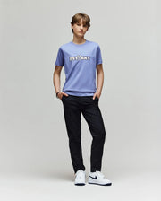 A person stands against a plain light gray background, wearing the Psycho Bunny KIDS WADE GRAPHIC TEE - KT0200055 in light purple, black pants, and white Nike sneakers, with hands in pockets and a relaxed posture.