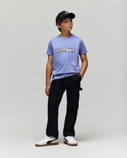 A boy in a black baseball cap, Psycho Bunny KIDS WADE GRAPHIC TEE - KT0200055 (light purple with "PSYBNY" and cartoon bunny), regular fit black pants, and white sneakers stands against a light gray background.