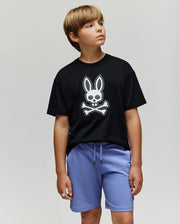 A boy with light brown hair wears the Psycho Bunny KIDS RYDER OVERSIZED FIT TEE (KT0200056) featuring an embroidered Bunny skull and crossbones, paired with light purple shorts. He stands against a plain light background, looking left.