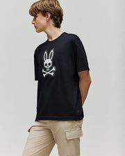 A person with short brown hair wears the Psycho Bunny KIDS RYDER OVERSIZED FIT TEE (KT0200056), featuring an embroidered Bunny skull and crossbones logo, paired with beige cargo pants, standing against a plain light background.