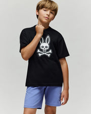 A boy with light brown hair stands confidently in the Psycho Bunny KIDS RYDER OVERSIZED FIT TEE (KT0200056), featuring an embroidered Bunny-skull logo, paired with light purple shorts against a plain gray background.