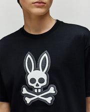 A person wearing the Psycho Bunny KIDS RYDER OVERSIZED FIT TEE (KT0200056), a black premium tee with soft Pima cotton and a large white rabbit head with crossbones graphic. Only the lower half of their face is visible.