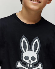 A child wears the Psycho Bunny KIDS RYDER OVERSIZED FIT TEE (KT0200056) in black, featuring a bold white skull and crossbones with bunny ears and teeth. Made of soft cotton, it highlights their lower face and upper torso.