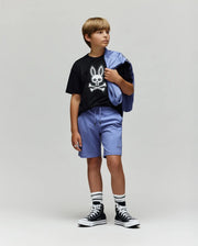 A boy stands against a plain background wearing the Psycho Bunny KIDS RYDER OVERSIZED FIT TEE (KT0200056), featuring an embroidered Bunny and crossbones, paired with light purple shorts, striped socks, black high-top sneakers, and a matching jacket.