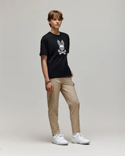 A young person with short hair stands against a plain background, wearing the Psycho Bunny KIDS RYDER OVERSIZED FIT TEE (KT0200056) featuring a white cartoon skull and bunny ears graphic, paired with beige pants and white sneakers.