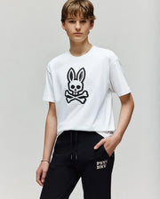 A person with short brown hair wears the Psycho Bunny KIDS RYDER OVERSIZED FIT TEE (KT0200056) with a black embroidered Bunny skull graphic, paired with black "PSY BNXY" pants, standing against a plain light background.