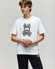 A young person stands against a plain background, wearing the Psycho Bunny KIDS RYDER OVERSIZED FIT TEE - KT0200056 with a large black bunny skull and crossbones graphic, paired with black pants. Their hands are loosely clasped in front.