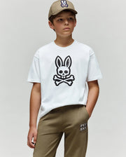 A boy stands against a plain background, wearing a khaki cap, khaki pants, and the Psycho Bunny KIDS RYDER OVERSIZED FIT TEE (KT0200056), featuring premium Pima cotton and an embroidered Bunny skull and crossbones graphic.
