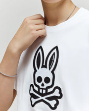 A person wears the kids' Ryder oversized fit tee (KT0200056) by Psycho Bunny, featuring a white base with a black skull and crossbones bunny design; their hand near their neck reveals a silver chain bracelet.