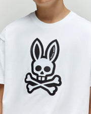 A person wearing the Psycho Bunny KIDS RYDER OVERSIZED FIT TEE (KT0200056), made of soft Pima cotton, featuring a large black cartoon bunny head with skull-like eyes and nose above crossed bones on the front.