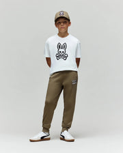 A young boy stands against a plain background, wearing the Psycho Bunny KIDS RYDER OVERSIZED FIT TEE - KT0200056 with a large bunny and crossbones graphic, olive green joggers, white sneakers, and an olive green cap with an embroidered Bunny logo.