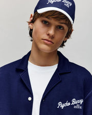 A young person in a navy Psycho Bunny cap and jacket wears the KIDS BASIL TAPED CAMP COLLAR SHIRT - KT0500007, a premium cotton-modal blend white shirt by Psycho Bunny, looking neutrally at the camera against a light plain background.