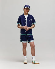 A person stands against a plain background wearing the Psycho Bunny KIDS BASIL TAPED CAMP COLLAR SHIRT (KT0500007) in a cotton-modal blend, with matching shorts, blue and white trim, a cap, white socks, and beige sneakers with green accents.