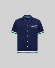 The Psycho Bunny KIDS BASIL TAPED CAMP COLLAR SHIRT (KT0500007) is a navy cotton-modal blend with white buttons, white and teal sleeve/hem trim, and "Psycho Bunny Est. 2005" embroidered in white on the left chest. Shown on gray background.