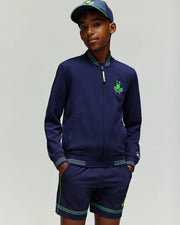 A young boy wears the Psycho Bunny RETAIL BALL CREW YOUTH JACKET (EN) - KT0500014 in navy blue with green accents and matching shorts, plus a navy blue cap, standing with hands in his pockets against a plain light background.