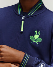 A person is wearing the Psycho Bunny RETAIL BALL CREW YOUTH JACKET (EN) - KT0500014: a navy jacket with green striped trim, a zipper, and a chest logo showing a rabbit head and crossed tennis rackets. Only their lower face, neck, and hand are visible.