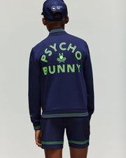 A person stands facing away, wearing the Psycho Bunny RETAIL BALL CREW YOUTH JACKET (EN) - KT0500014 in navy with bold neon "PSYCHO BUNNY" lettering and bunny logo, paired with navy shorts and a matching cap for a sporty, casual look.