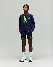 A young boy models the Psycho Bunny RETAIL BALL CREW YOUTH JACKET (KT0500014) in navy blue, paired with matching shorts, neon green graphic tee, white socks, and sneakers against a plain light gray background.
