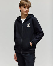 A young person stands with one hand in their pocket against a light background, wearing the Psycho Bunny KIDS WADE CORD PATCH ZIP HOODIE (KT0500016) in black, featuring a small bunny and crossbones chest design.