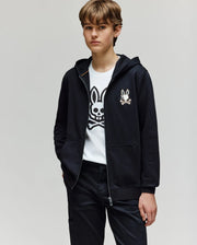 A young person with short brown hair wears the Psycho Bunny KIDS WADE CORD PATCH ZIP HOODIE (KT0500016) over a white T-shirt with a cartoon bunny skull logo, standing with one hand in their pocket against a plain light background.