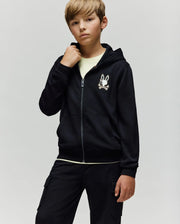 A blond-haired boy wears the Psycho Bunny KIDS WADE CORD PATCH ZIP HOODIE (KT0500016), featuring an embroidered hood and a white cartoon chest graphic, over a light tee and black pants, against a plain light background.
